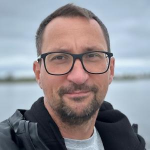 Andreas Beger outdoors on an overcast day, in black-framed glasses and a dark jacket, with a lake and low grey cloud behind him.